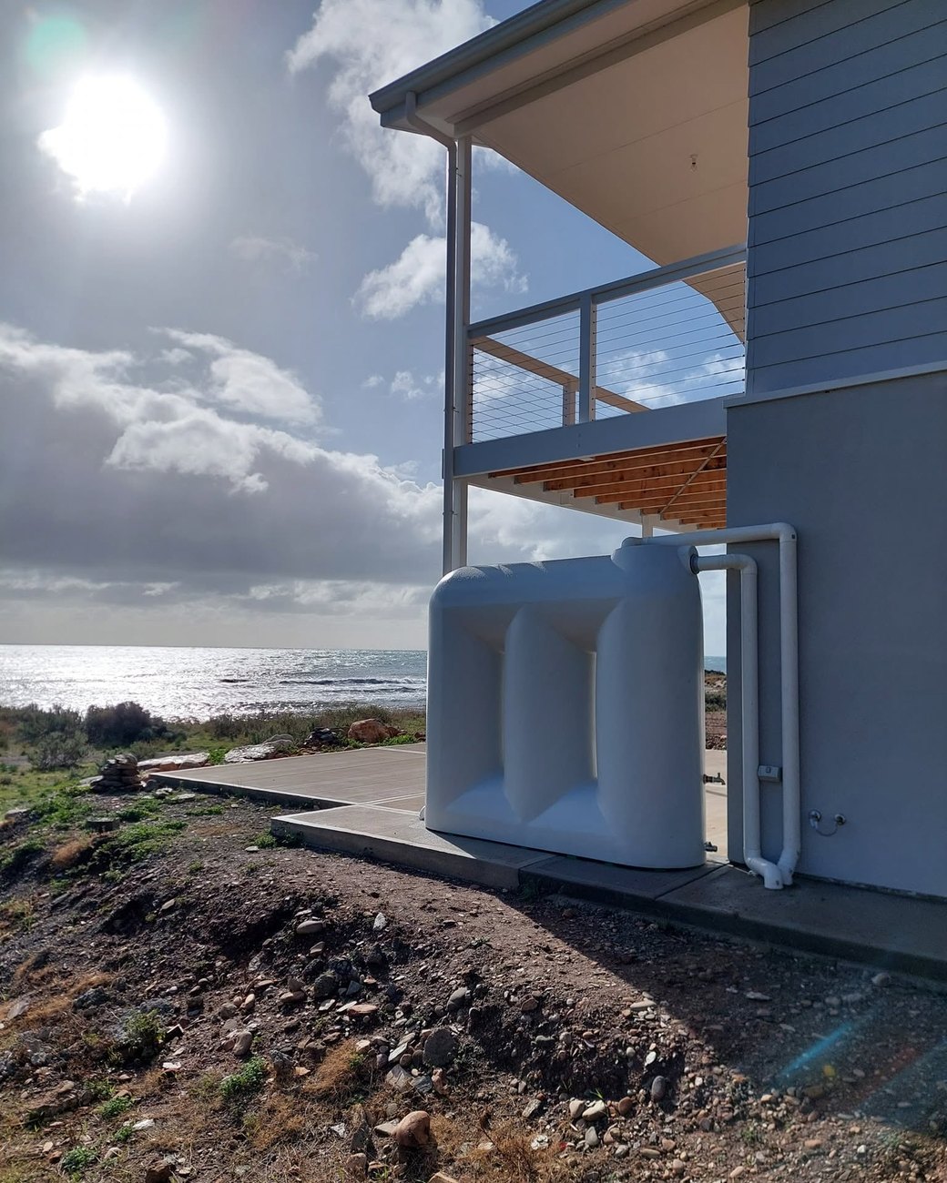 A slimline rainwater tank plumbed into the downpipe of a coastal house, with the sea behind